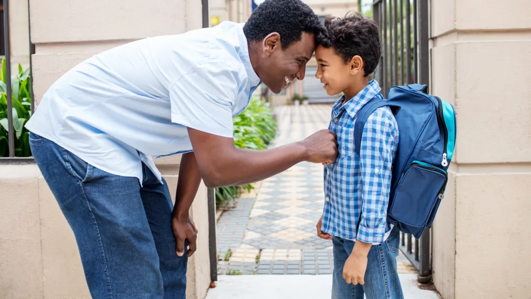 A father and a son standing outside,. The son is wearing a backpack and the father is bending over, comforting him.