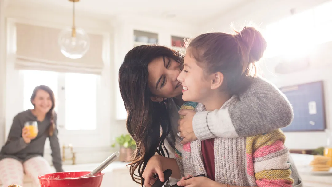 A family of three women are in the kitchen, one daughter hugging the youngest while she flips pancakes.