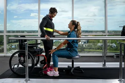 A woman standing up out of a wheelchair with the help of a healthcare worker.
