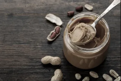 A spoonful of peanut butter sitting on a jar of peanut butter and peanuts surrounding the jar on a table.