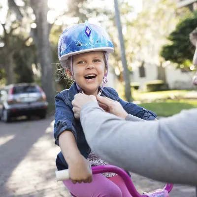 A father tightens the helmet strap of his young daughter on her pink bicycle.