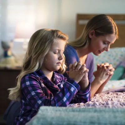 A young girl prays by her bedside with her mother before bed.
