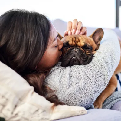 Woman laying on a couch hugging and kissing her fawn colored french bulldog