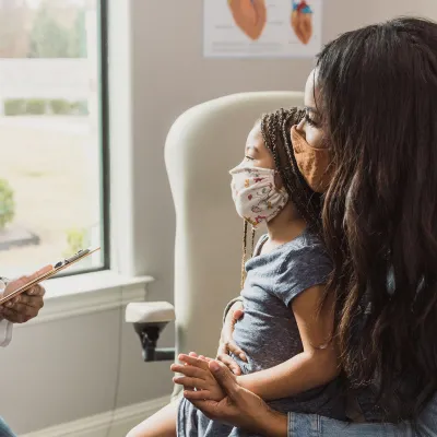 Mother and Child Visit Their Pediatrician in an Exam Room in Front of a Large Window While Wearing Face Masks.