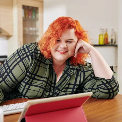 A Woman Smiles as She Sits at Her Kitchen Table Viewing Content on Her Tablet.