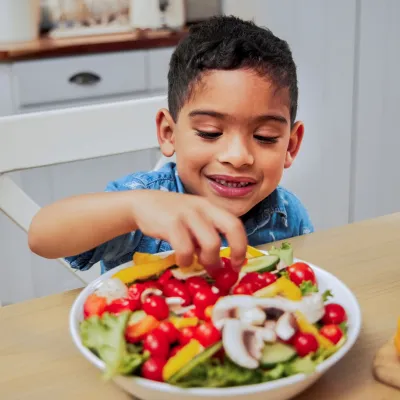 A Young Boy Grabs Some Vegetables From a Salad on a Kitchen Table.
