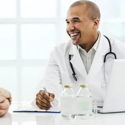 A senior man visits his male doctor for his annual screening. Both are seated at a table.