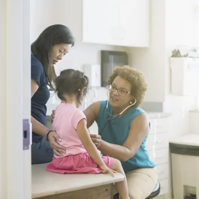 A female AHS doctor listens to a little girl's heart in the exam room while her mother comforts her