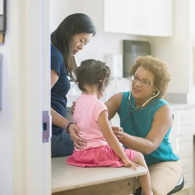 A female AHS doctor listens to a little girl's heart in the exam room while her mother comforts her