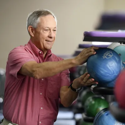 An adult man crouches to choose a bowling ball at the bowling alley.