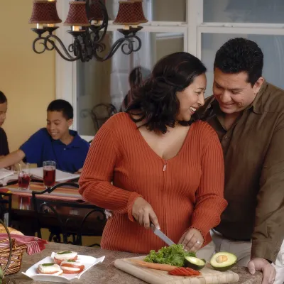 A couple shares a sweet moment while the wife prepares dinner, as their kids do homework in the background