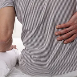 A man holds his lower back while sitting on a bed. 