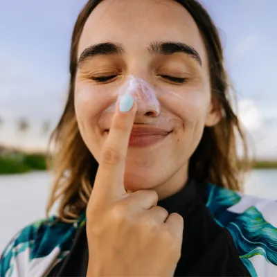 A woman on a beach wearing sunscreen on her nose and touching her nose with her index finger.