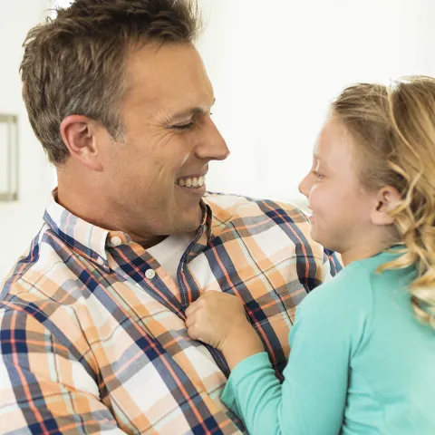 A Caucasian father smiles at his young daughter, holding her in his arms.