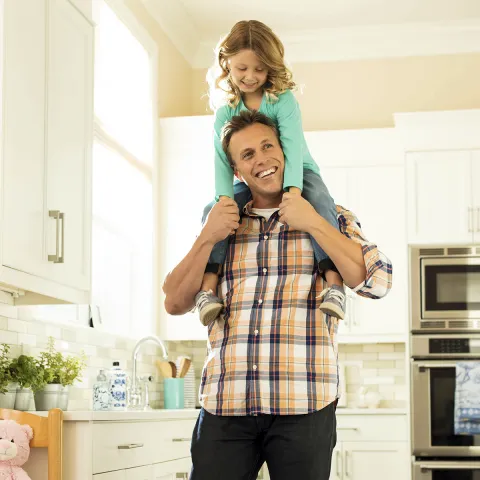 A Caucasian father carries his young daughter on his shoulders in their home.