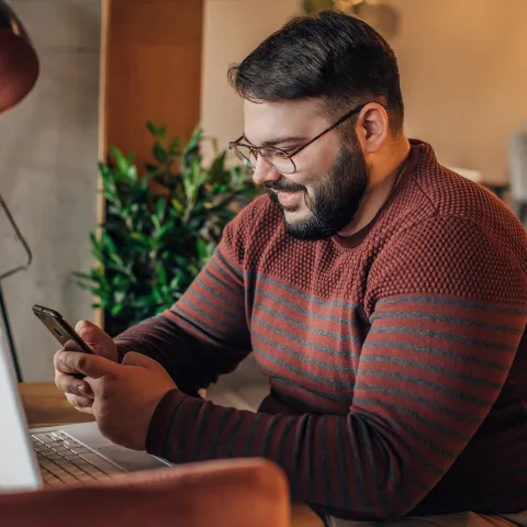 A Man Smiles as He Uses a Cellphone While Sitting at a Desk in front of a Laptop.