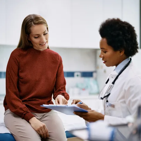A Doctor Speaks to Her Patient in an Exam Room While Going Over Her Charts on a Laptop.