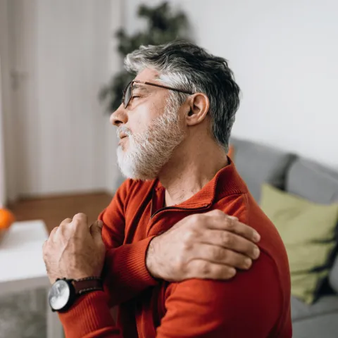 An Older Gentleman Rubs a Sore Shoulder While Sitting on His Sofa at Home
