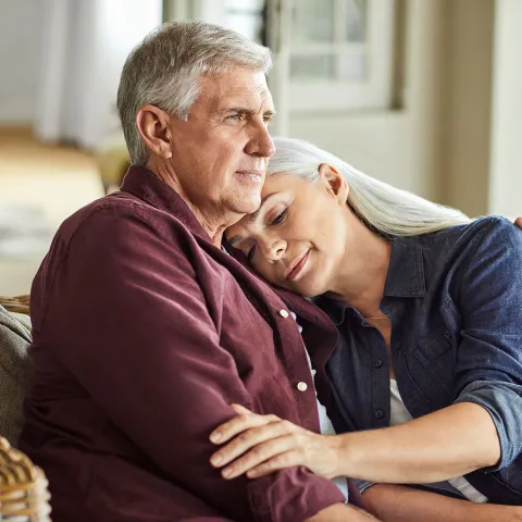 A Senior Woman Rests Her Head on Her Senior Husbands Shoulder While They Sit On a Couch at Home