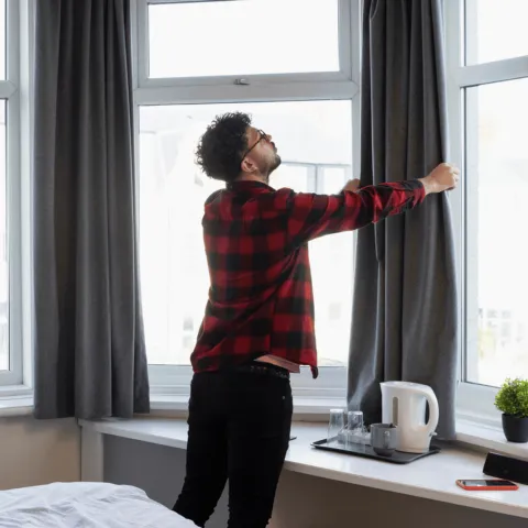 A man in a red and black checkered shirt is opening gray curtains in a room with large windows.