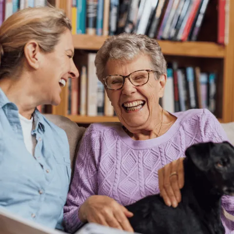 Two Women Laughing While Sitting On A Couch With A Black Dog In Front Of A Bookshelf