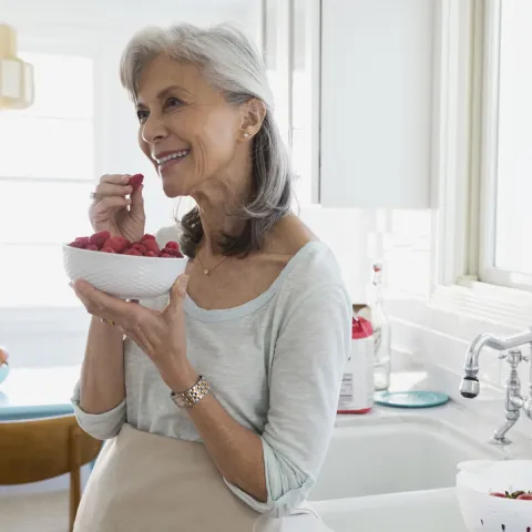An older woman eats raspberries from a bowl in her kitchen.