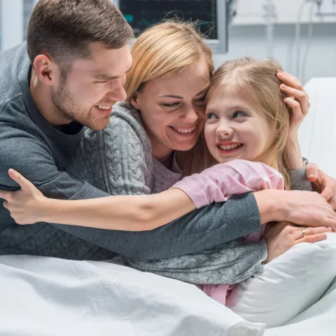 A family hugging their young daughter in her hospital bed.