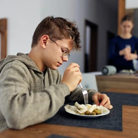 A boy sits at a dining room table eating lunch in the foreground while his mother is in the background adding seasoning to food on a plate.
