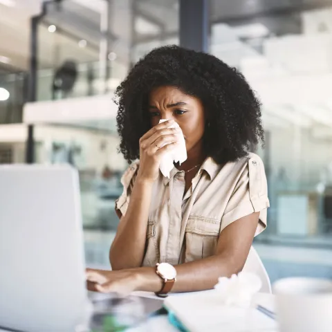 A woman blowing her nose, looking at a laptop.