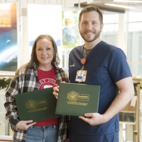 A white woman with brown hair and wearing blue jeans, a red tee shirt, and a black and white plaid button up shirt stands with a white, lightly bearded man with short brown hair who is wearing navy blue scrubs. They are both holding green leather folders that read "DAISY Award for Extraordinary Nurses"