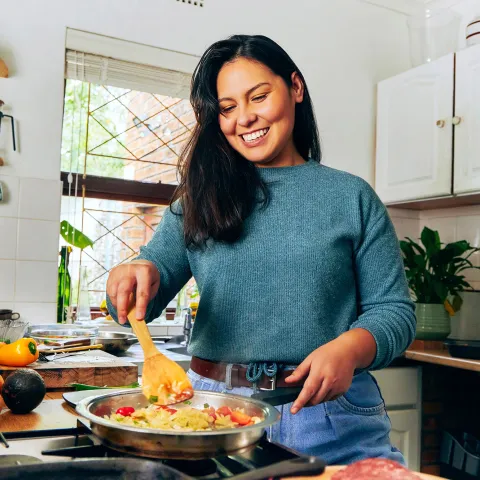 Smiling person stirs vegetables in a pan in a bright, cluttered kitchen.