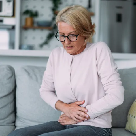 A senior woman sitting on a couch holding her stomach in discomfort while at home.