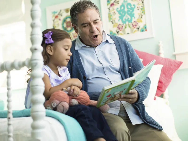 A grandfather reads a children's book to his granddaughter in her bedroom.