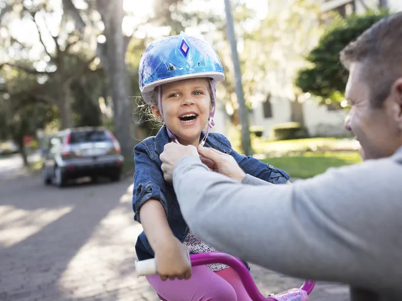 A father tightens the helmet strap of his young daughter on her pink bicycle.