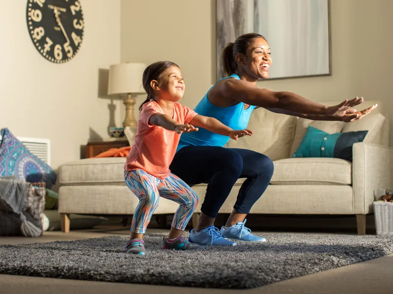 A mother and her young daughter exercise together in the living room.