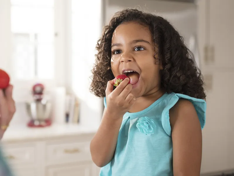 A young African American girl eats strawberries in the kitchen.