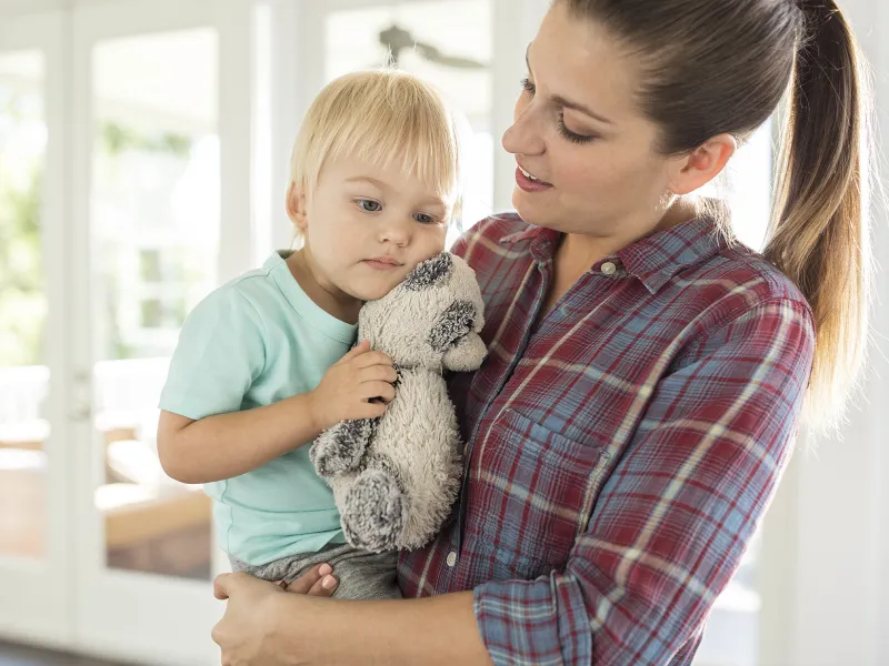 A Caucasian mother holds her young in the living room son with his stuffed animal. 