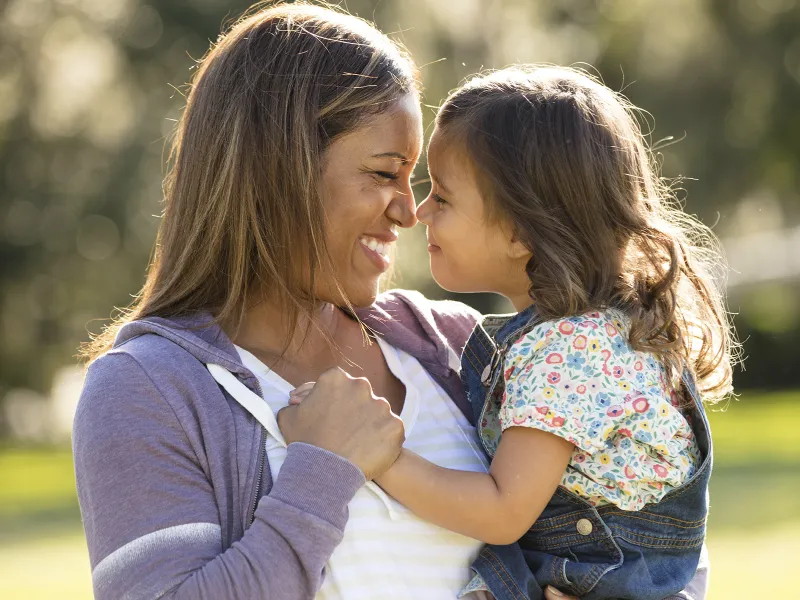 A mother smiles at her young daughter while holding her. 