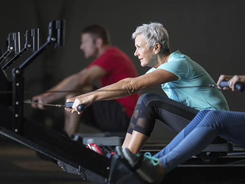 An elderly Caucasian woman exercises on a rowing machine at the gym.
