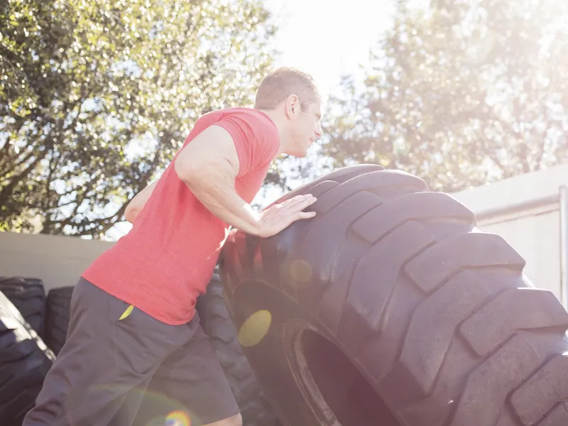 A young Caucasian man exercises outdoors by flipping large tires.