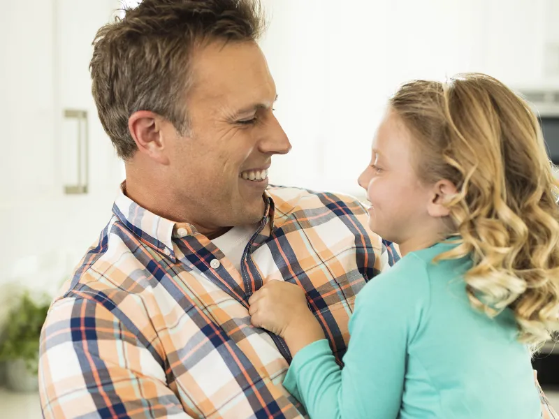 A Caucasian father smiles at his young daughter, holding her in his arms.