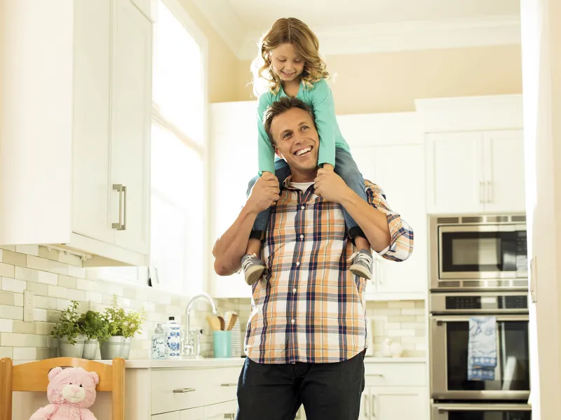 A Caucasian father carries his young daughter on his shoulders in their home.