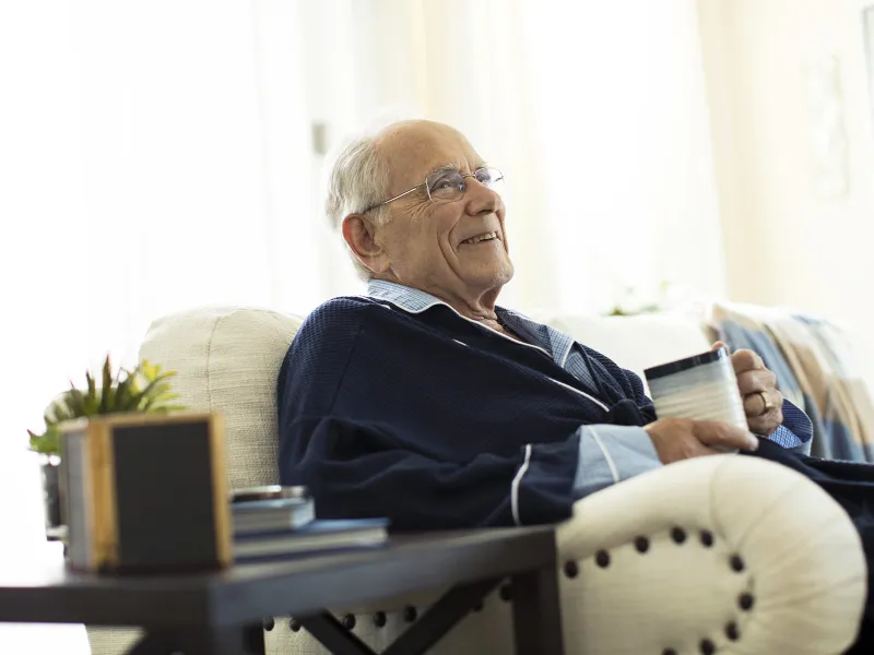 An elderly Caucasian man sits on the sofa with a mug of coffee