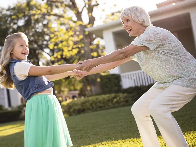 A Caucasian grandmother and granddaughter spin around and dance in a yard.