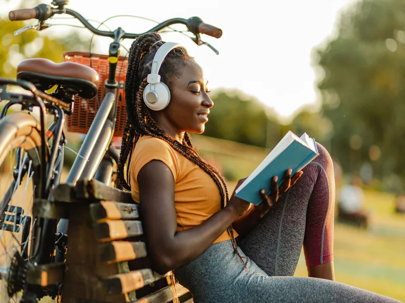 A Woman Takes a Break From Riding Her Bike By Sitting on a Park Bench and Reading a Book.
