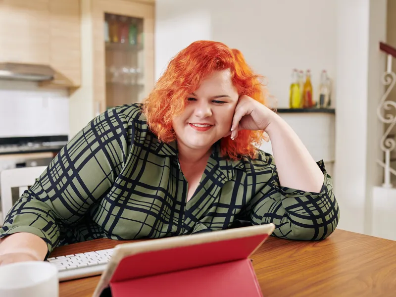 A Woman Smiles as She Sits at Her Kitchen Table Viewing Content on Her Tablet.