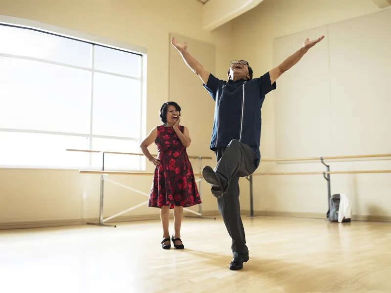 A Hispanic man leaps for joy in a dance studio while his wife smiles on.
