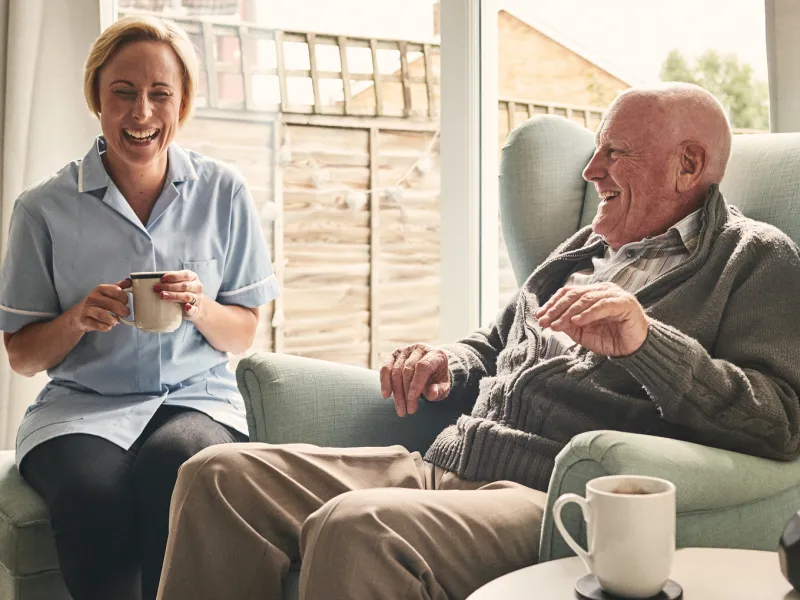 A senior man, reclined in a chair, laughs with his caretaker, who is seated next to him.
