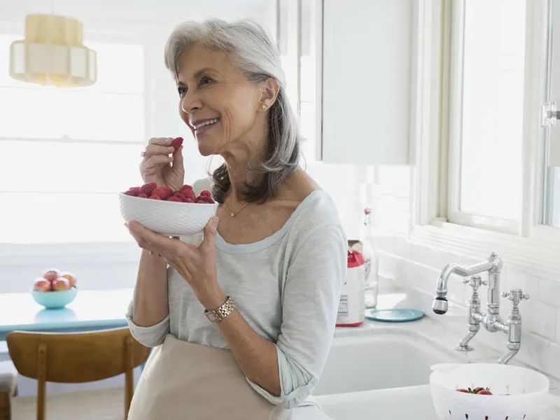 An older woman eats raspberries from a bowl in her kitchen.