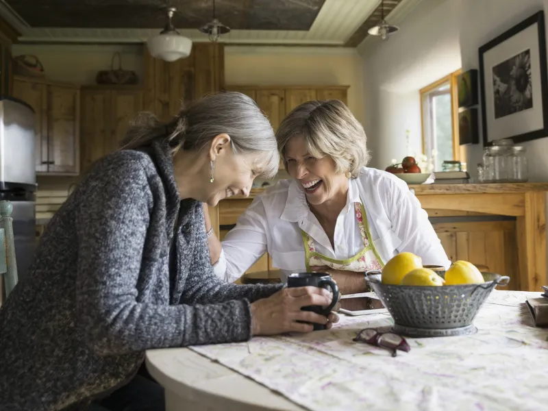 Two adult Caucasian women laugh together at a dining room table.